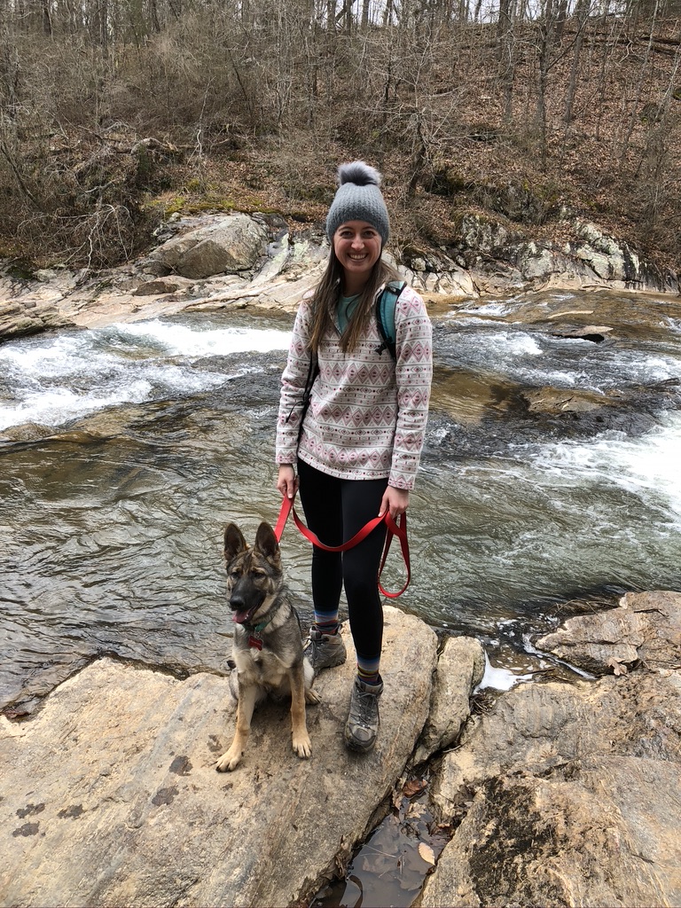 Girl standing next to dog on a leash in front of a creek. Hiking while tent camping with dogs.