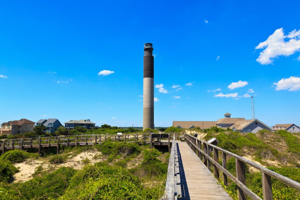 Tall, skinny lighthouse at Oak Island right beside local houses and a boardwalk.