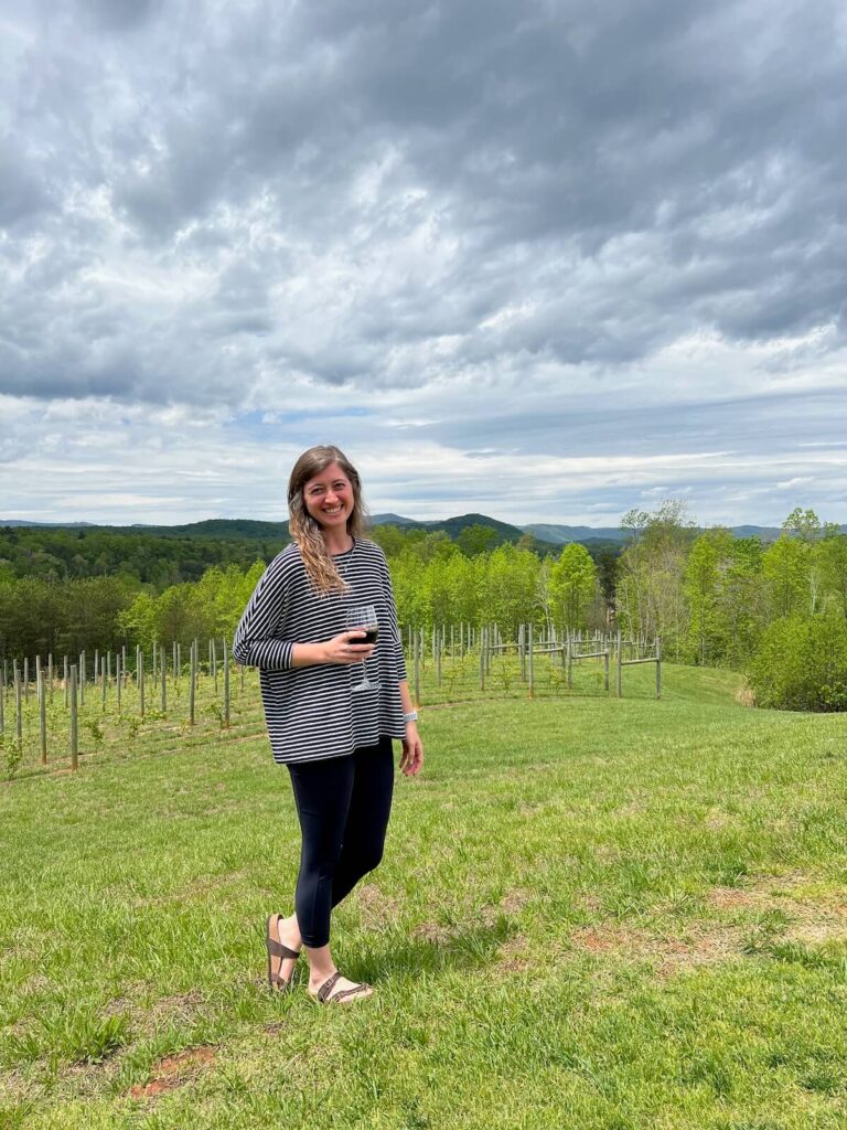 Girl standing on a hill in front of a vineyard holding a glass of wine.