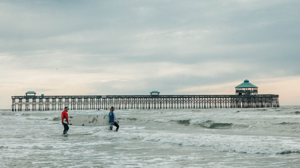 Two surfers carrying a board into the ocean with a pier in the background.