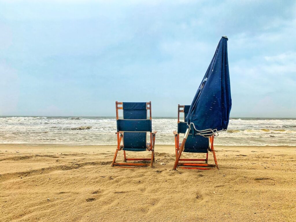 Two beach chairs facing the ocean with a closed, leaning umbrella behind them.