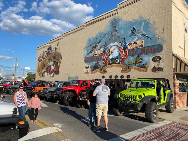 Jeeps lined up in front of Veterans of Boaz mural and people walking by.