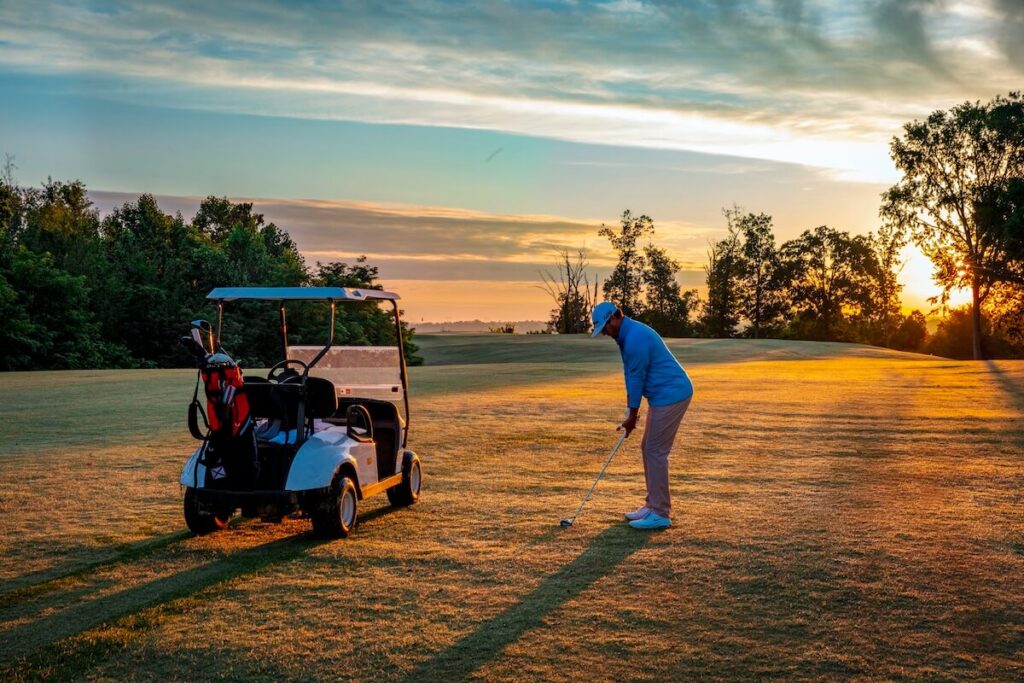 Man on golf course with golf cart about to hit a ball.