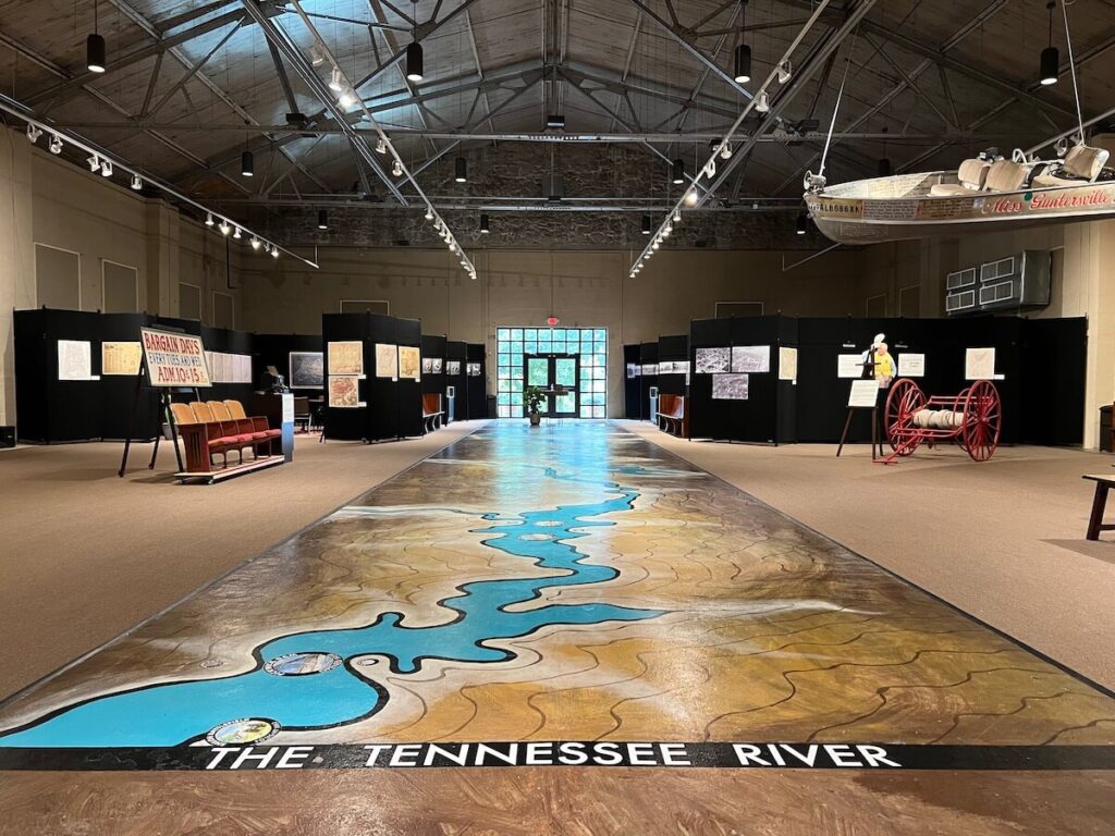 Mural of the TN River on the floor, boat hanging from the ceiling, and exhibits in the back of a museum room.