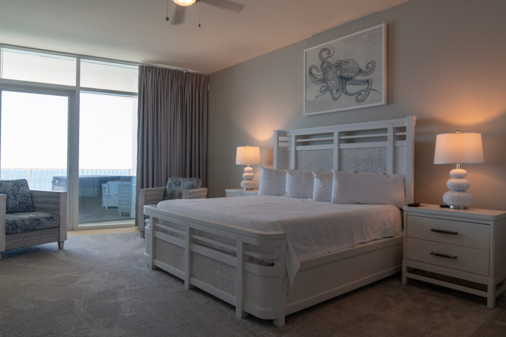 Master bedroom in a condo at the beach with a glass door leading to the balcony and a white color scheme. Pitching to hotels is a great way to secure these type of stays.