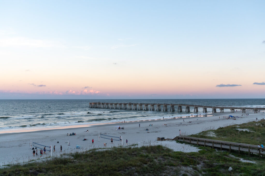 A long, concrete pier jutting out into the ocean at Jacksonville Beach with lots of people out on the beach. I was able to come here because I learned how to work with tourism boards.