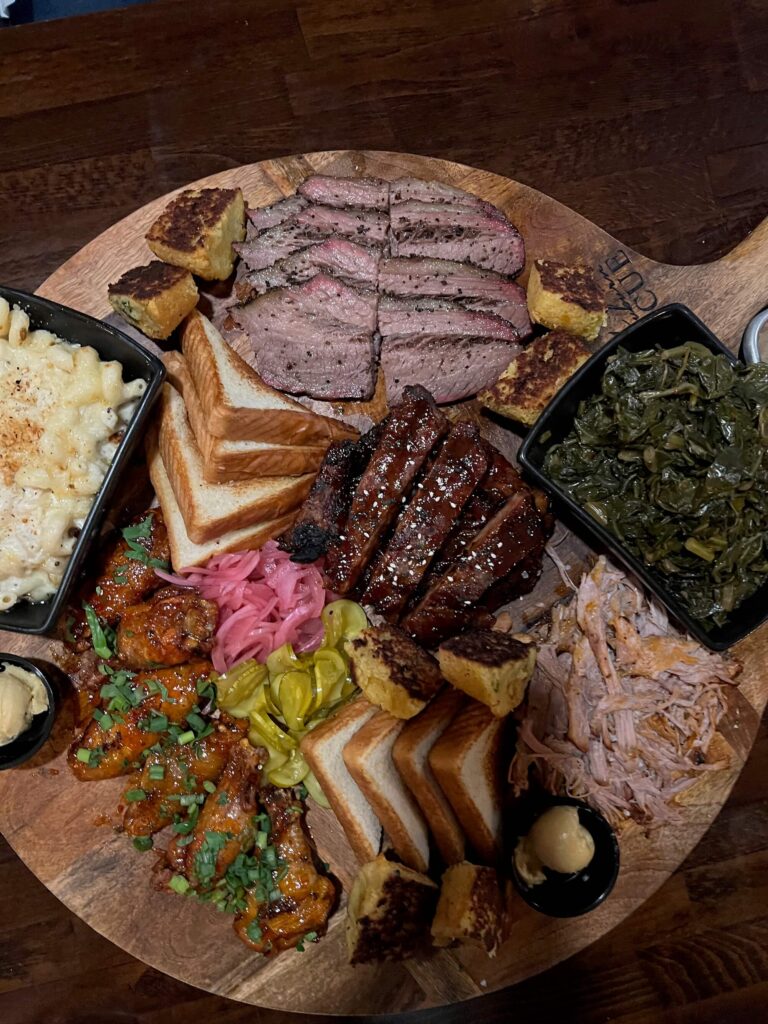 cutting board full of ribs, wings, BBQ, brisket, mac & cheese, collards, cornbread, and toast.