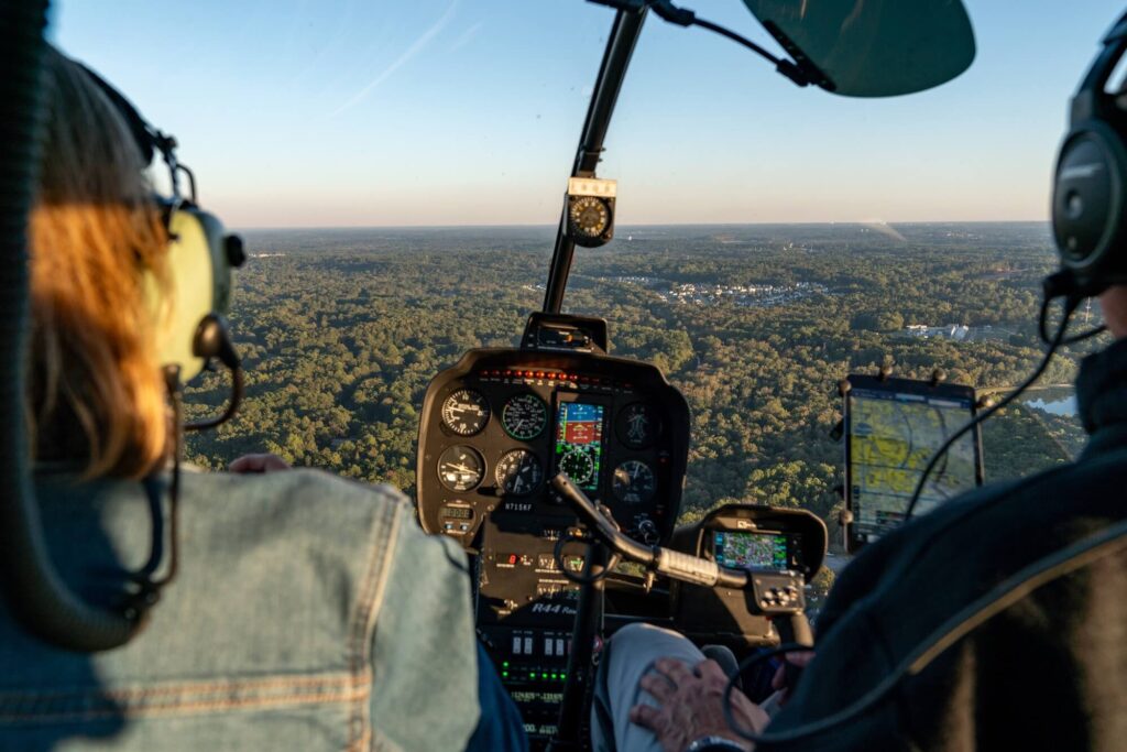 view out the front of a helicopter between two people showing wooded area below with a small collection of buildings
