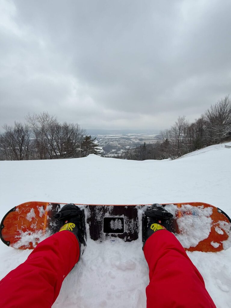person's legs and feet strapped into a snowboard looking out at the valley from the top of a ski hill
