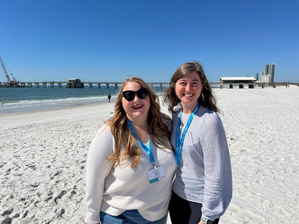 Two ladies on the beach posing for the camera while attending a media marketplace or a trade show.