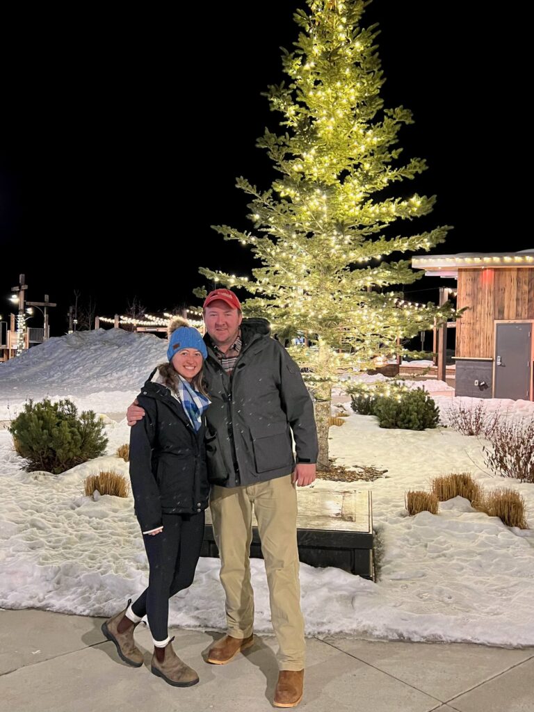 Man and woman posing in front of tree with Christmas lights dressed in cold weather gear.