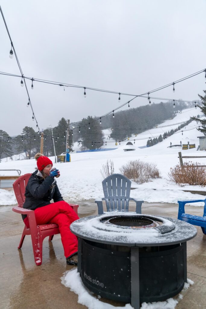 Girl sitting by unlit fire pit at the base of a ski mountain while sipping coffee in her snowboarding gear.